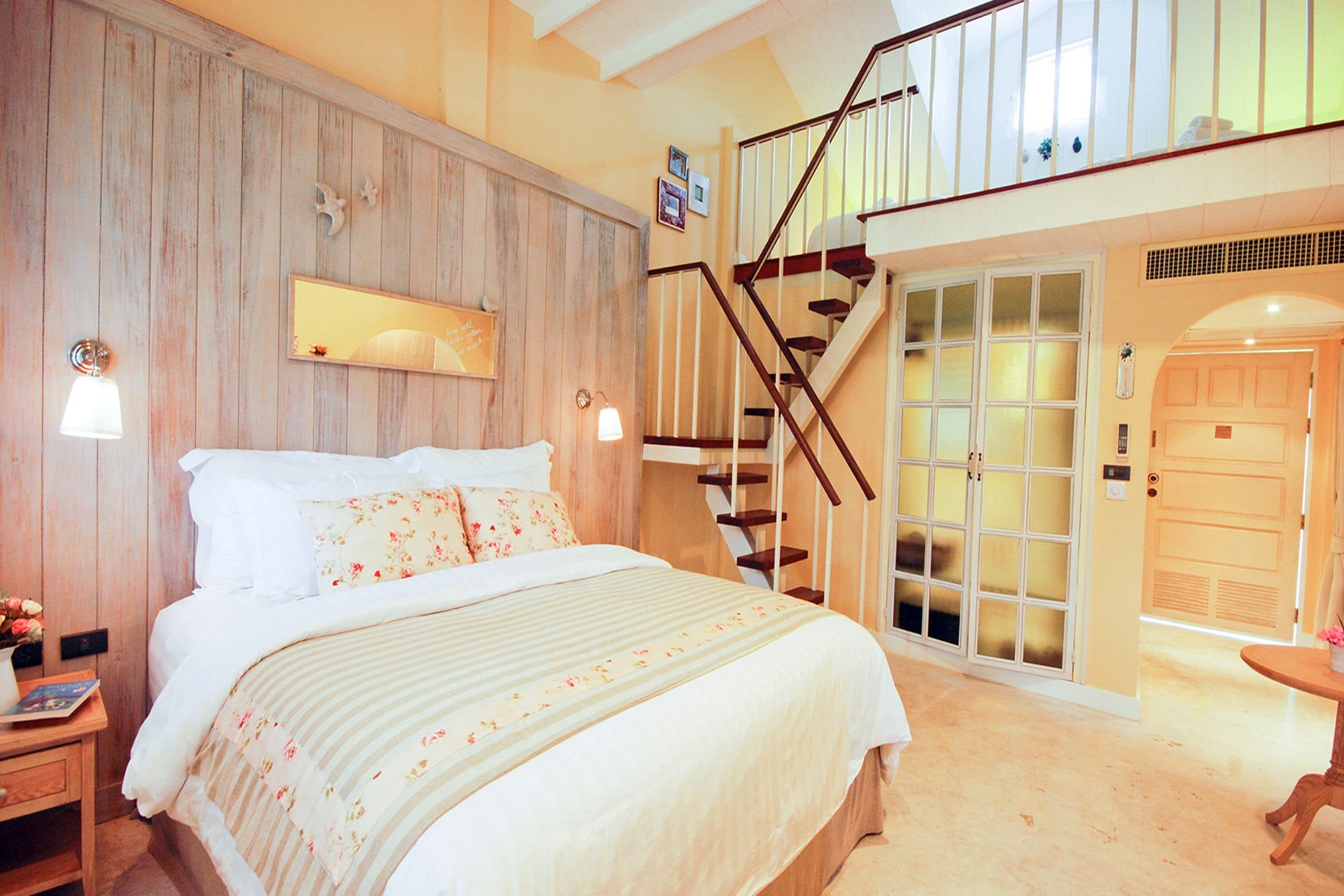 A bedroom with a large bed, wooden paneling on the wall, a staircase, and a door leading to another room.