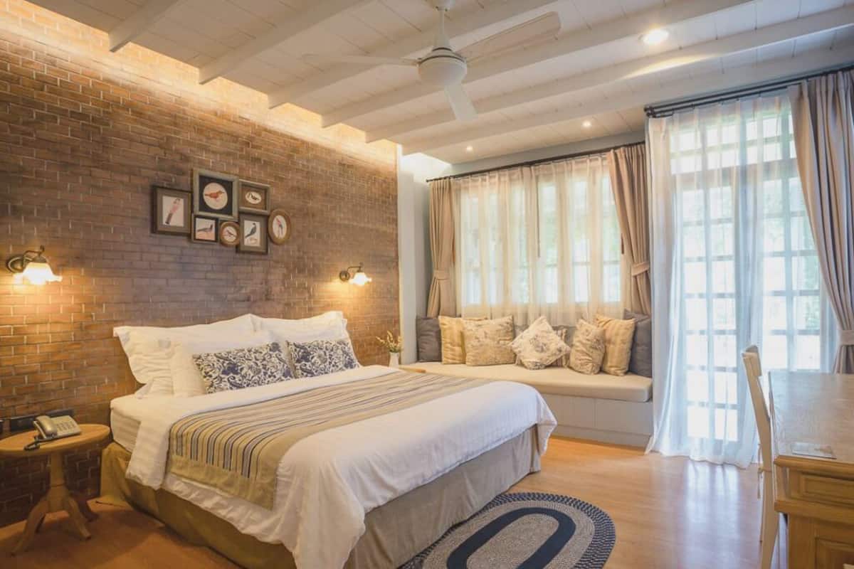 Une chambre avec un mur en briques, un lit avec de nombreux oreillers, une table de chevet avec une lampe et une fenêtre avec des rideaux.