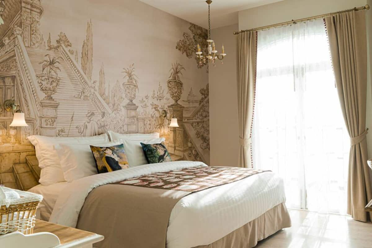 Une chambre spacieuse avec un grand lit, un lustre et une vue sur l’extérieur à travers de fines rideaux.