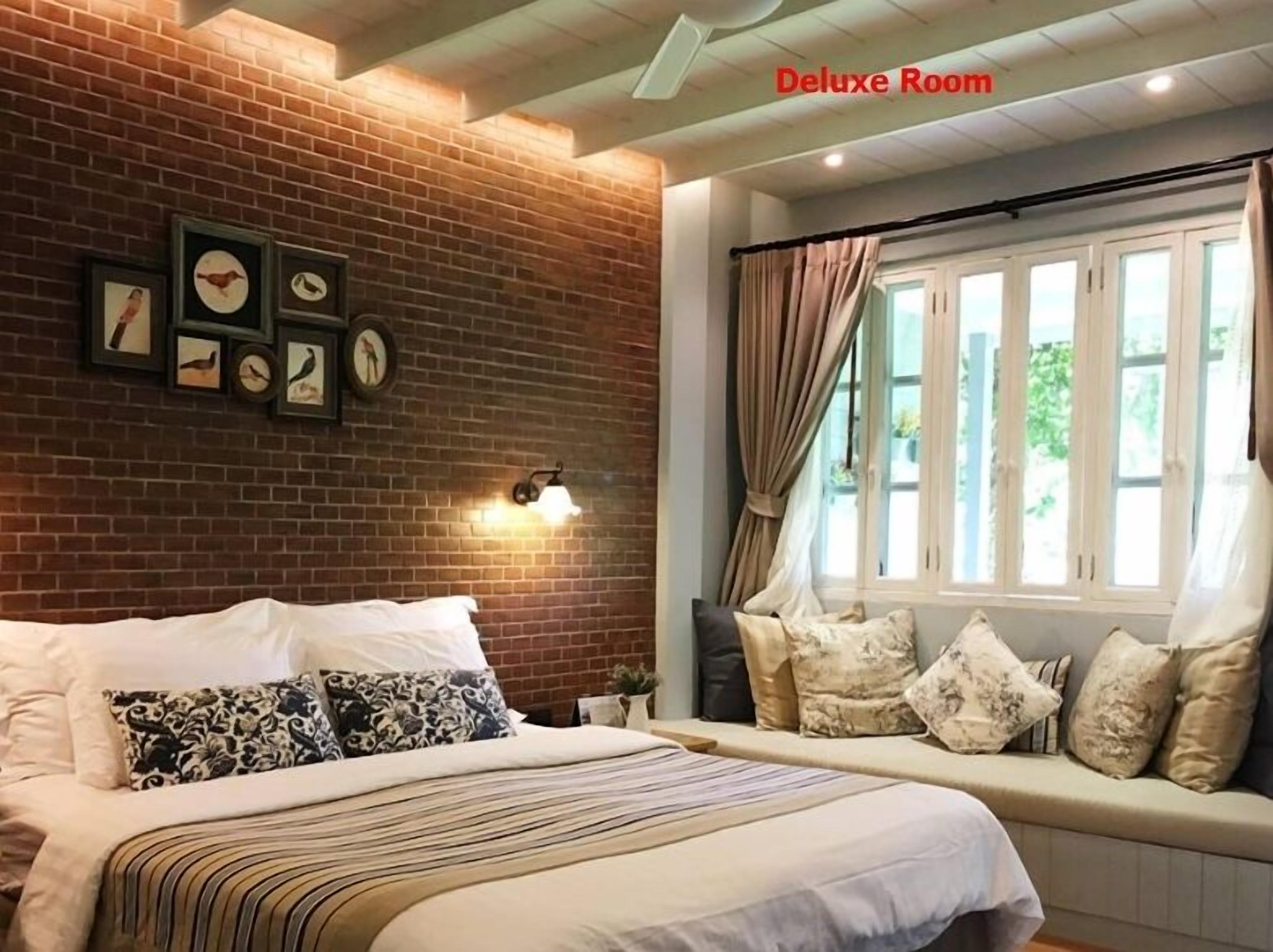 A room with a bed, a bench with cushions, a brick wall, framed pictures, and a window with curtains.
