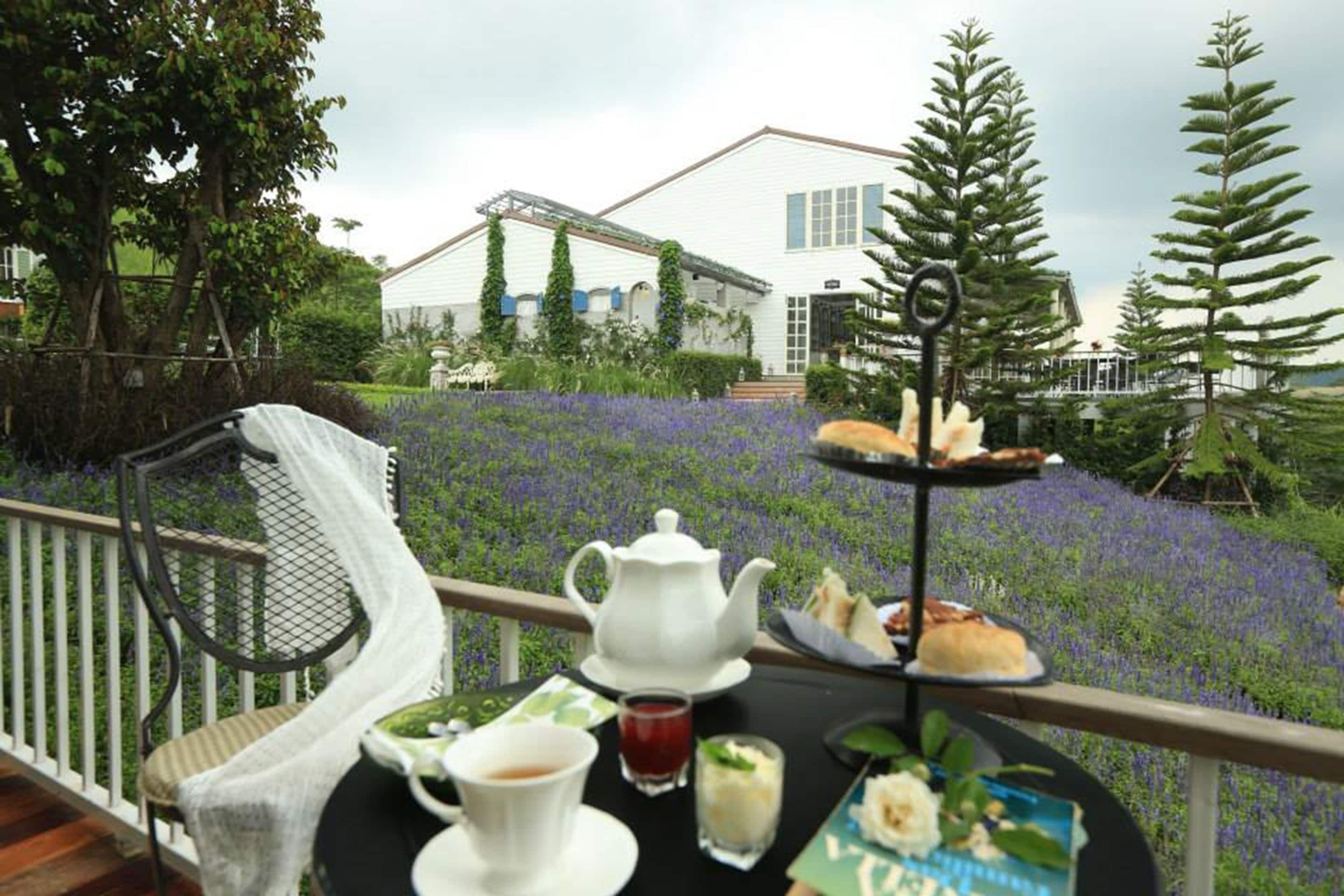 A table set with a teapot, cups, and a dessert tray on a balcony overlooking a garden with purple flowers and a white building in the background.