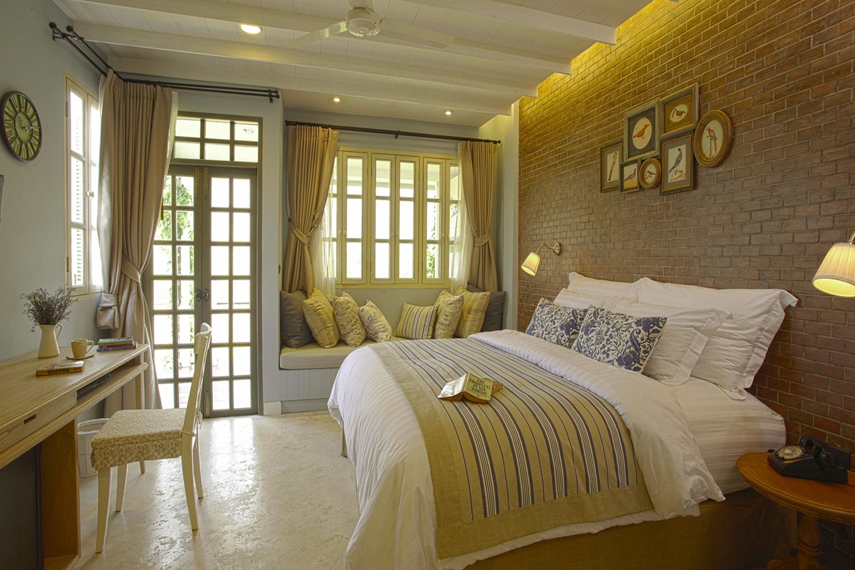 A bedroom with a large bed, a desk with a chair, a window with curtains, and a brick wall with framed pictures.