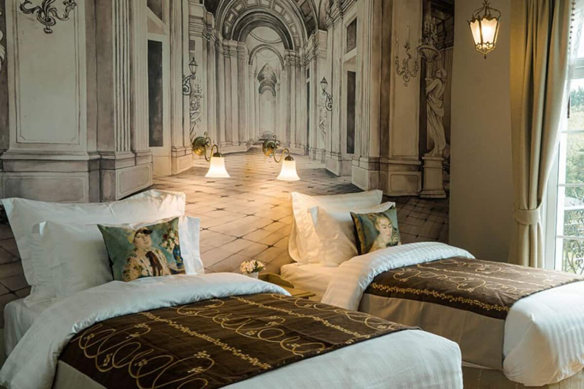 Une chambre d’hôtel avec deux lits, des colonnes richement décorées et un lustre.