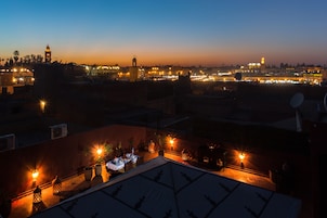 Una terraza en la azotea con mesas y sillas, rodeada de edificios y luces de la ciudad al anochecer.