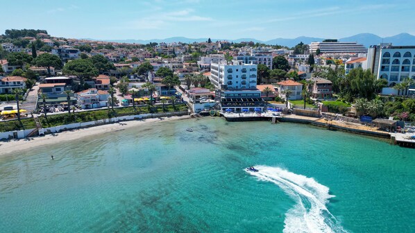 Een kustplaats met een jachthaven, helder turquoise water en diverse gebouwen, waaronder een opvallend wit hotel met balkons.