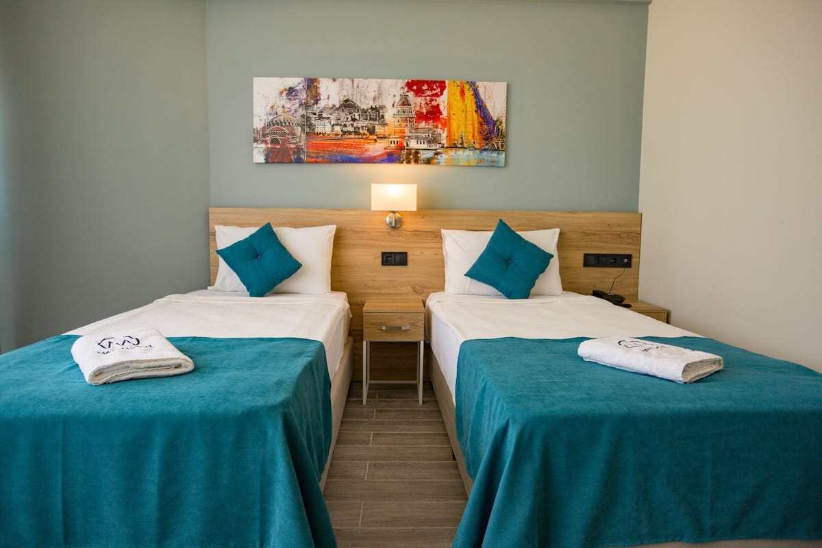 Une chambre d’hôtel avec deux lits, chacun recouvert d’une couvre-lit turquoise et agrémenté d’oreillers blancs. Au-dessus des lits, on peut voir un tableau. Il y a également une table de chevet avec une lampe et le sol est en bois.