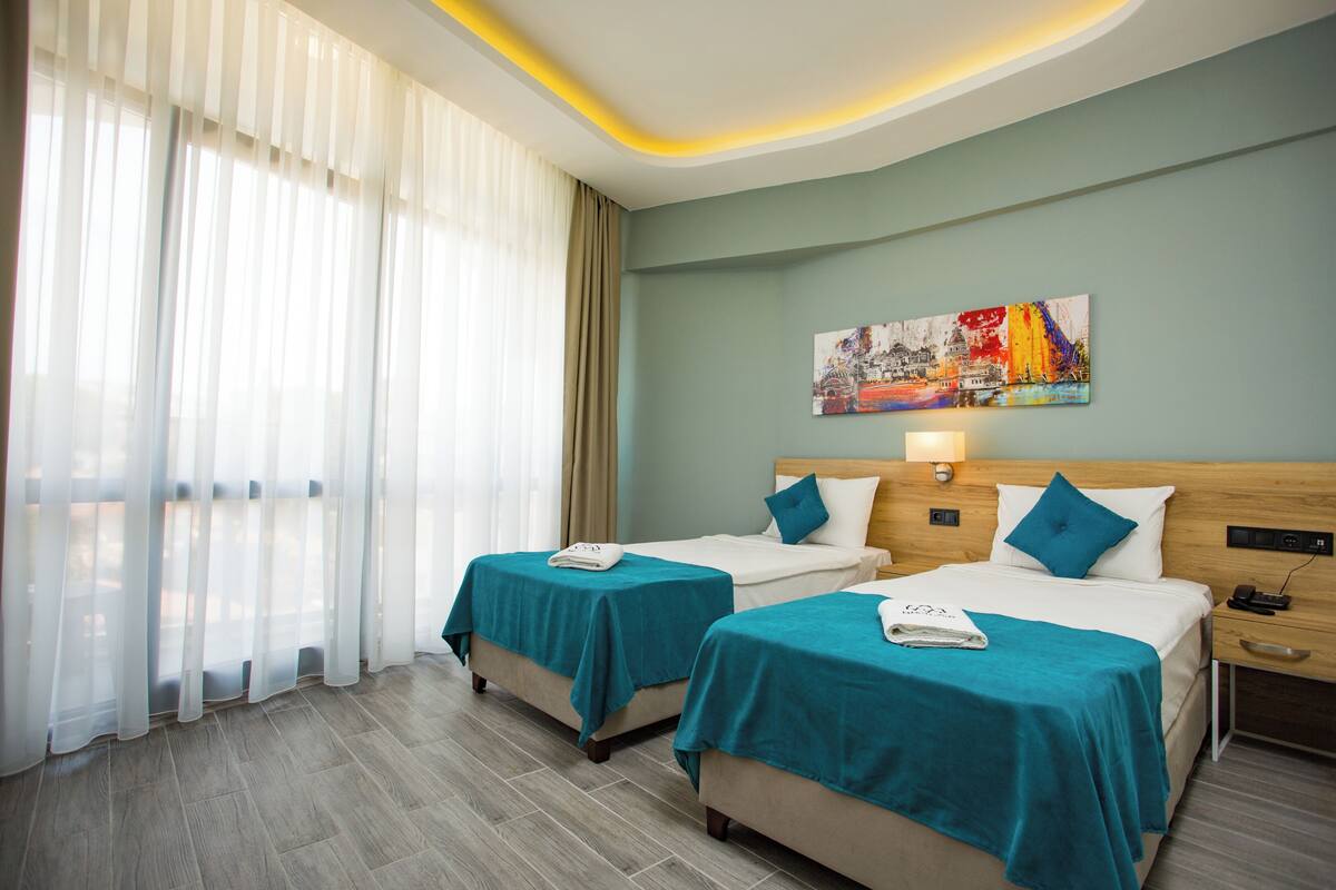 Une chambre d’hôtel avec deux lits, une grande fenêtre avec des rideaux légers, un tableau au mur et une tête de lit en bois.