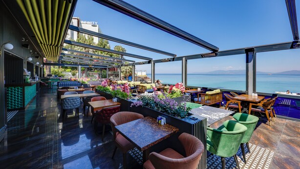 An outdoor seating area with tables and chairs, a pool, and a view of the sea. - Marti Beach Hotel (Kusadasi)