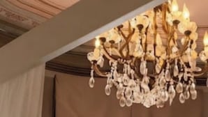 Une pièce dotée d’un lustre, de grandes fenêtres avec des rideaux, d’un fauteuil avec une couverture, et offrant une vue sur les bâtiments environnants.