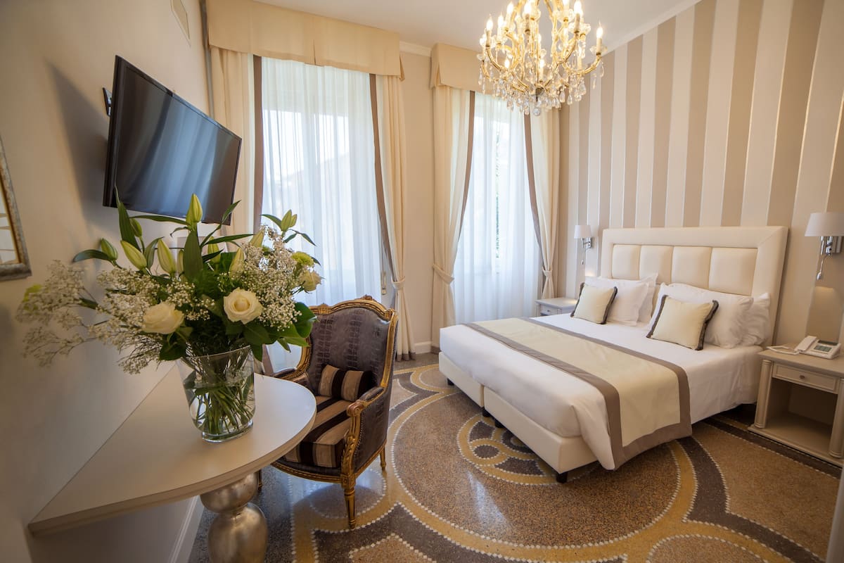 Une chambre d’hôtel avec un lit, un téléviseur fixé au mur, un vase avec des fleurs, une chaise et un sol recouvert de moquette.