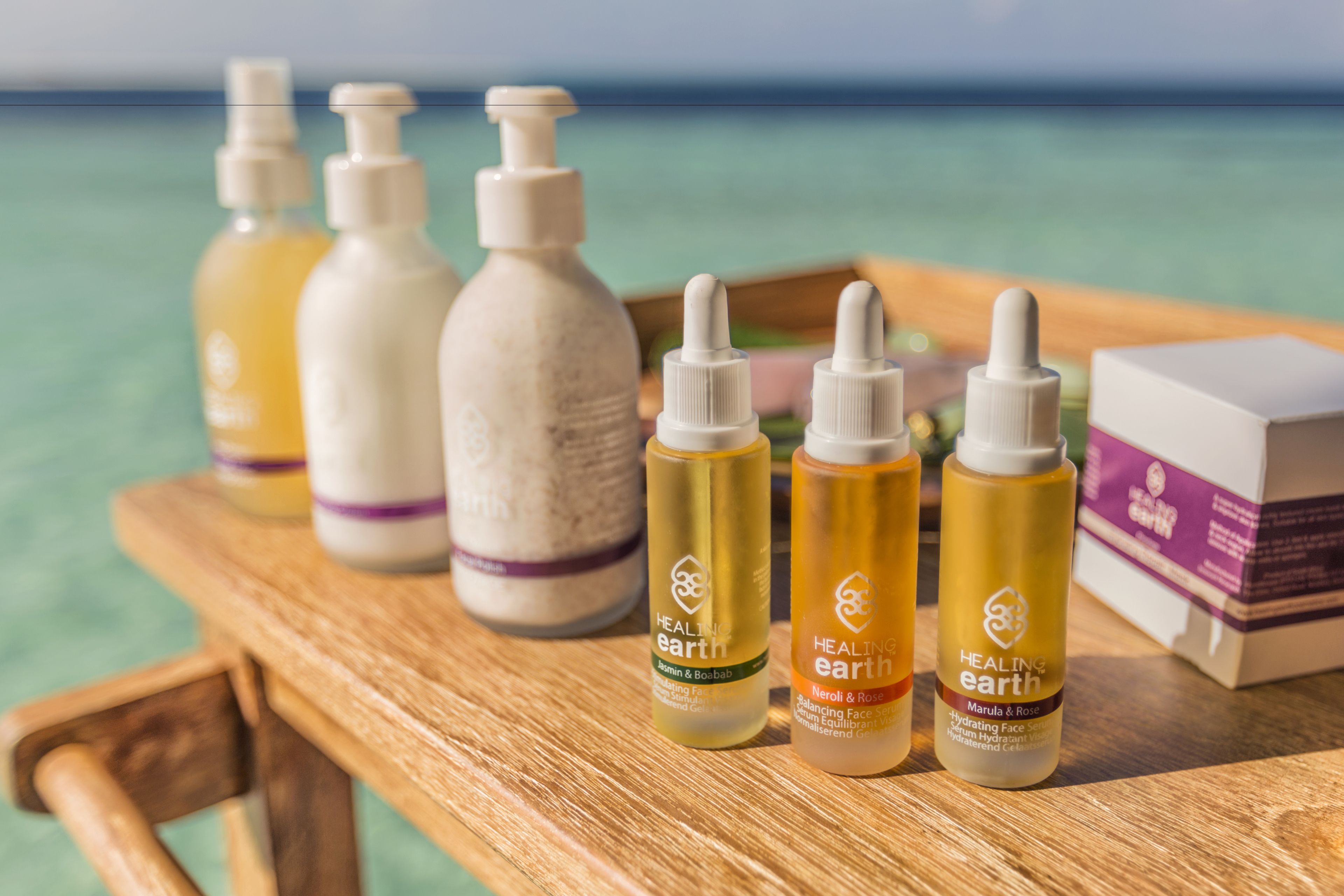 A wooden table with various skincare products from 'Healing Earth' brand, including bottles and a box, set against a backdrop of a beach and ocean.