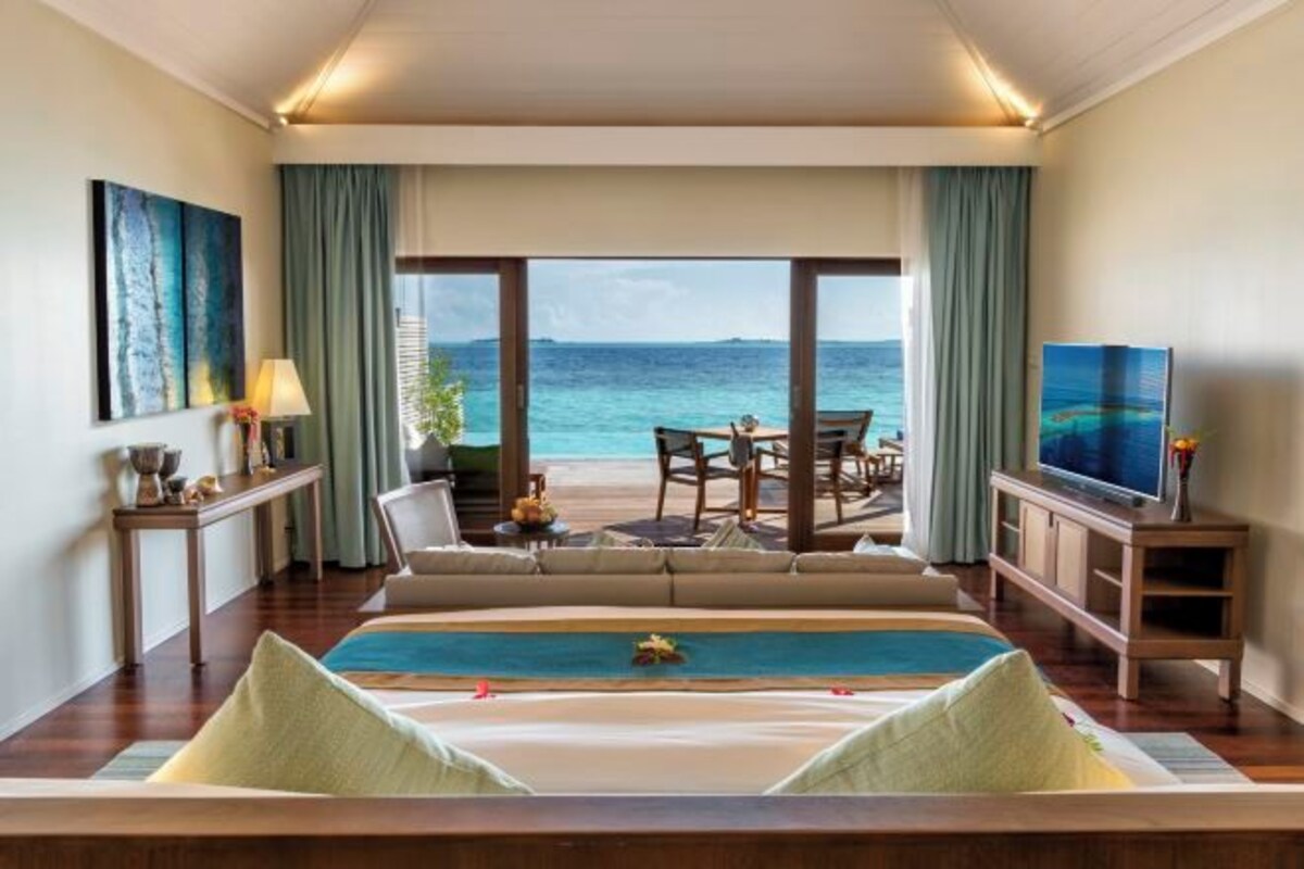 A hotel room with a large bed, a TV, a dining area with a view of the sea, and a balcony with a table and chairs.