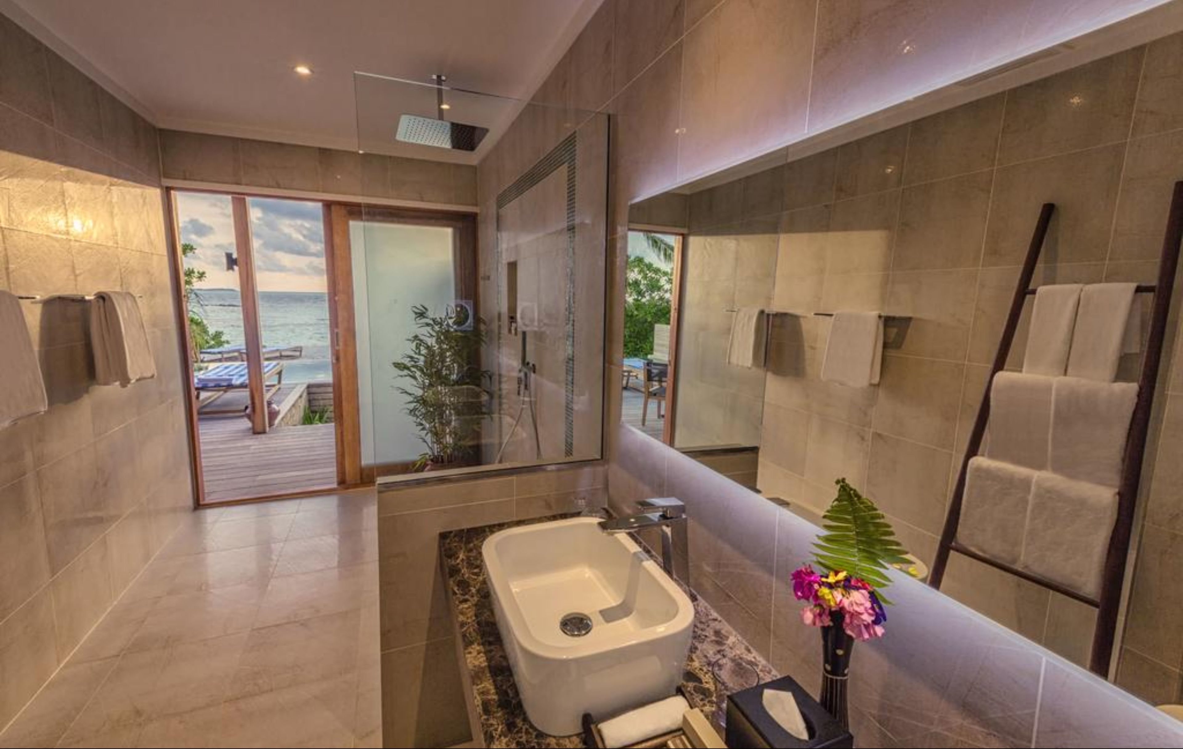 A modern bathroom with a large bathtub, a shower, and a glass-enclosed door leading to a balcony with a view of the sea.