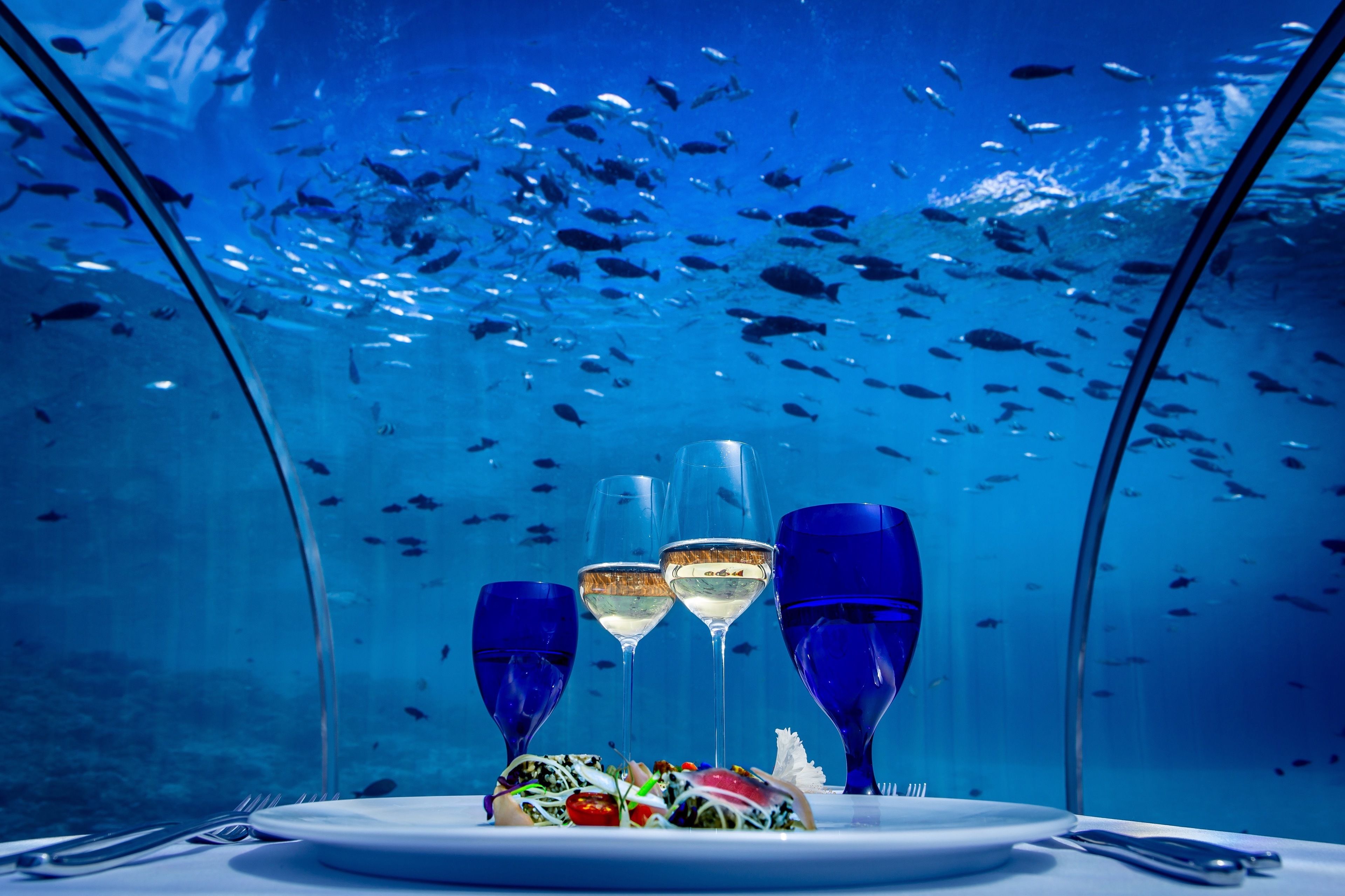 Un espacio comedor submarino con una mesa preparada para dos, rodeado por una estructura de vidrio.
