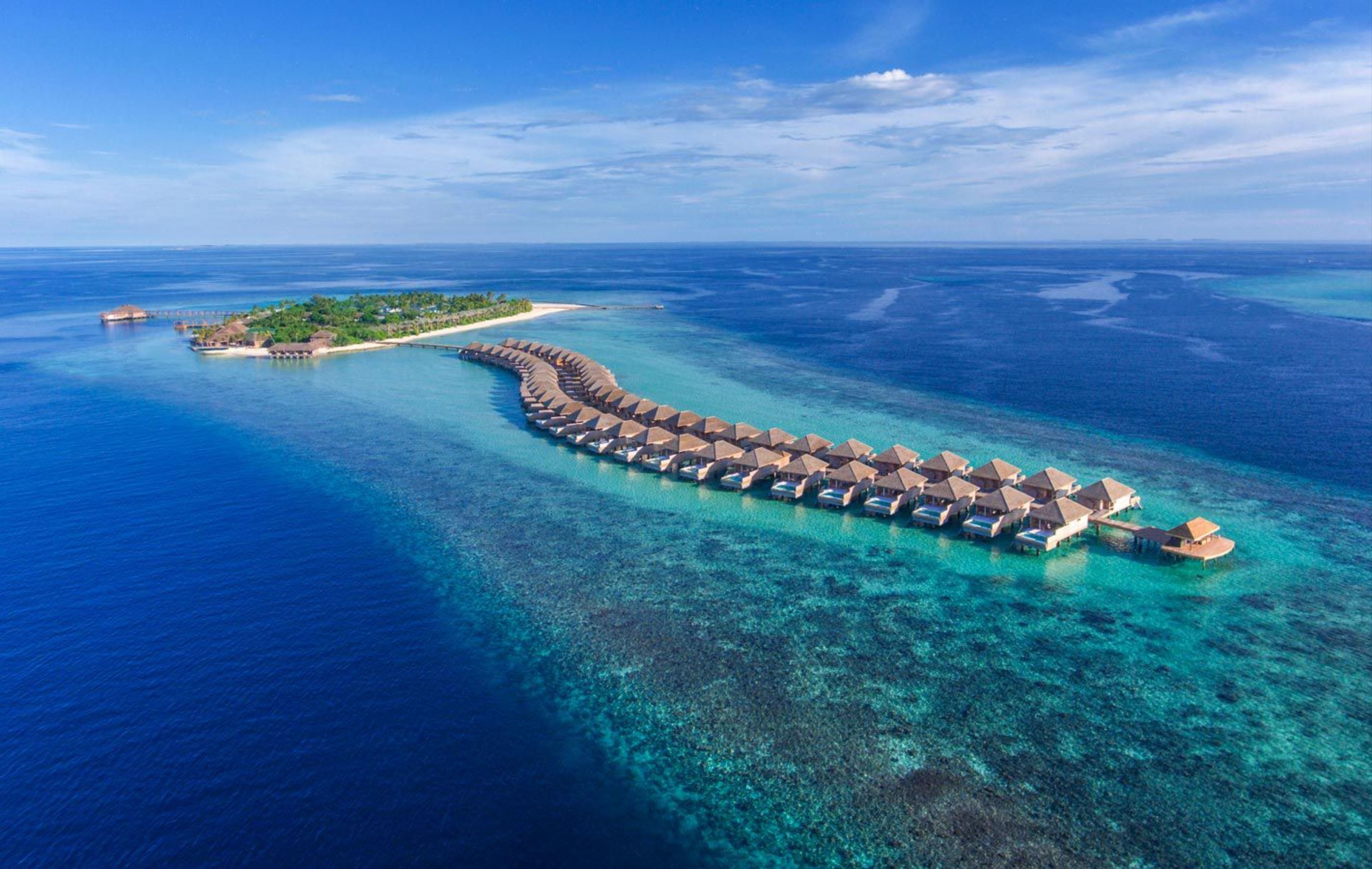 An overwater bungalow resort with a continuous row of thatched-roof bungalows connected by a walkway, surrounded by clear turquoise waters and a small island with greenery.