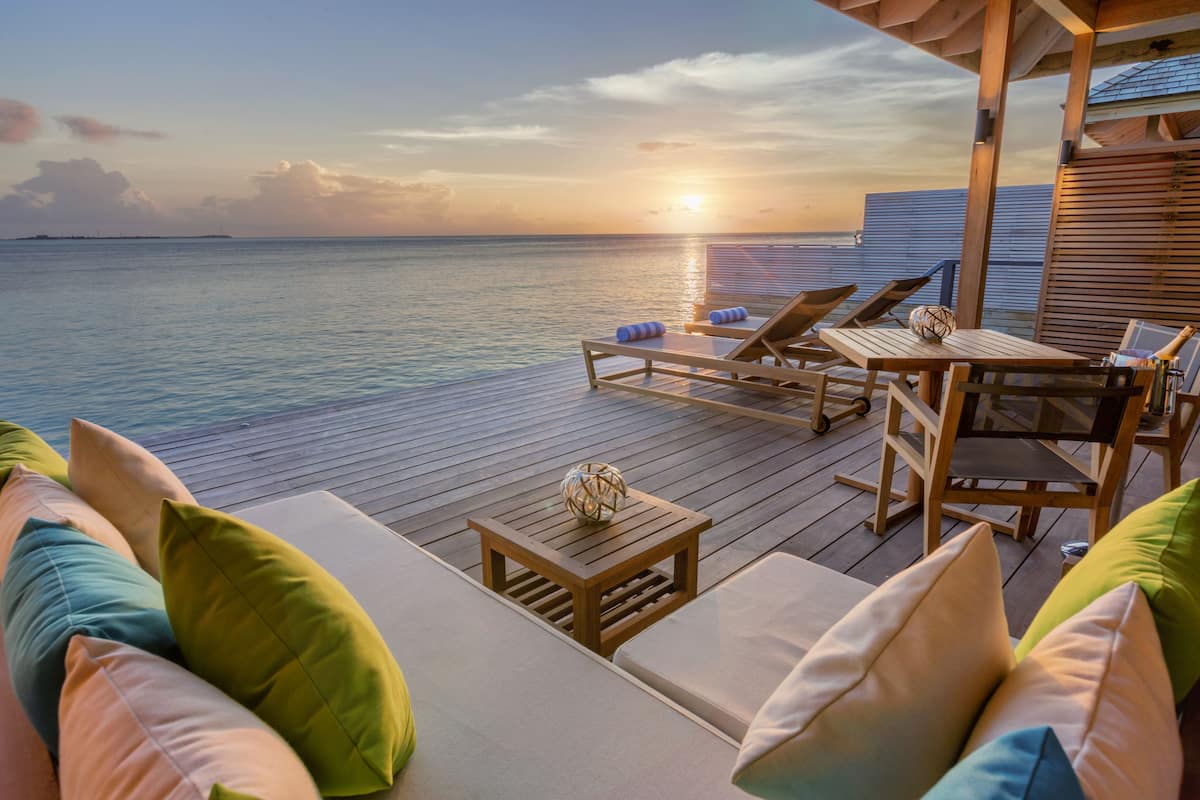Een houten terras met lounge fauteuils en een tafel, met uitzicht op een rustige zee bij zonsondergang.