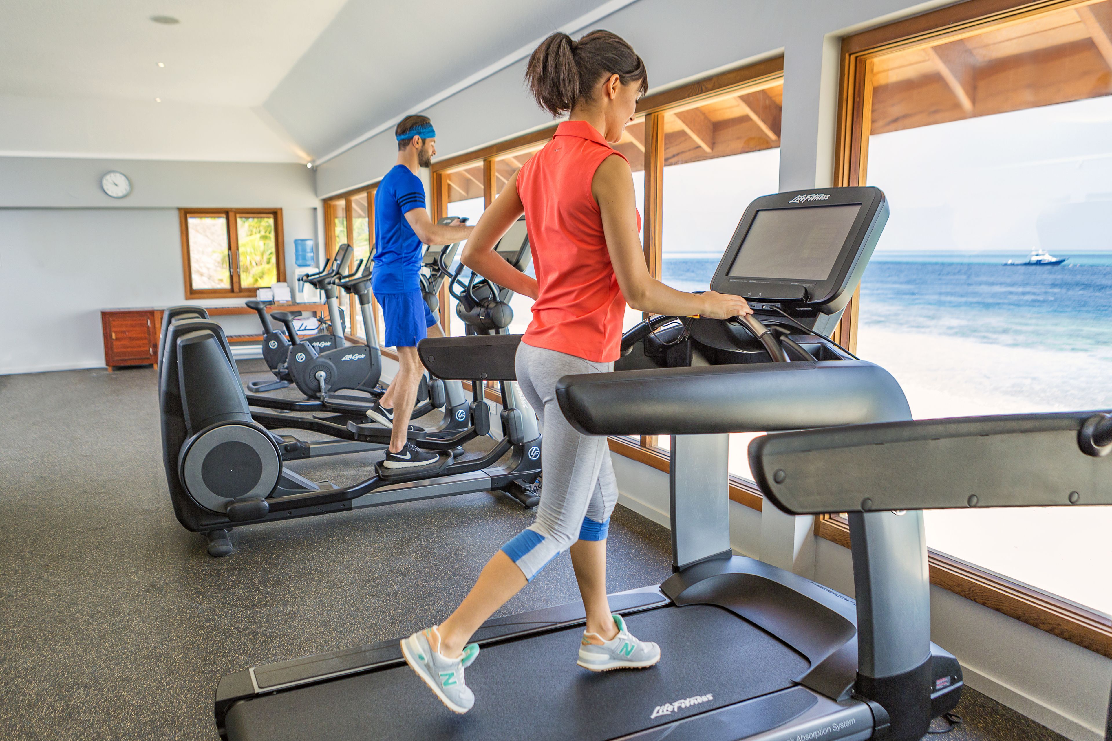 Un gimnasio con cintas de correr y equipo de ejercicio, amplias ventanas con vista al mar y un reloj en la pared.