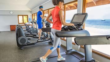 Un gimnasio con cintas de correr y equipo de ejercicio, amplias ventanas con vista al mar y un reloj en la pared.