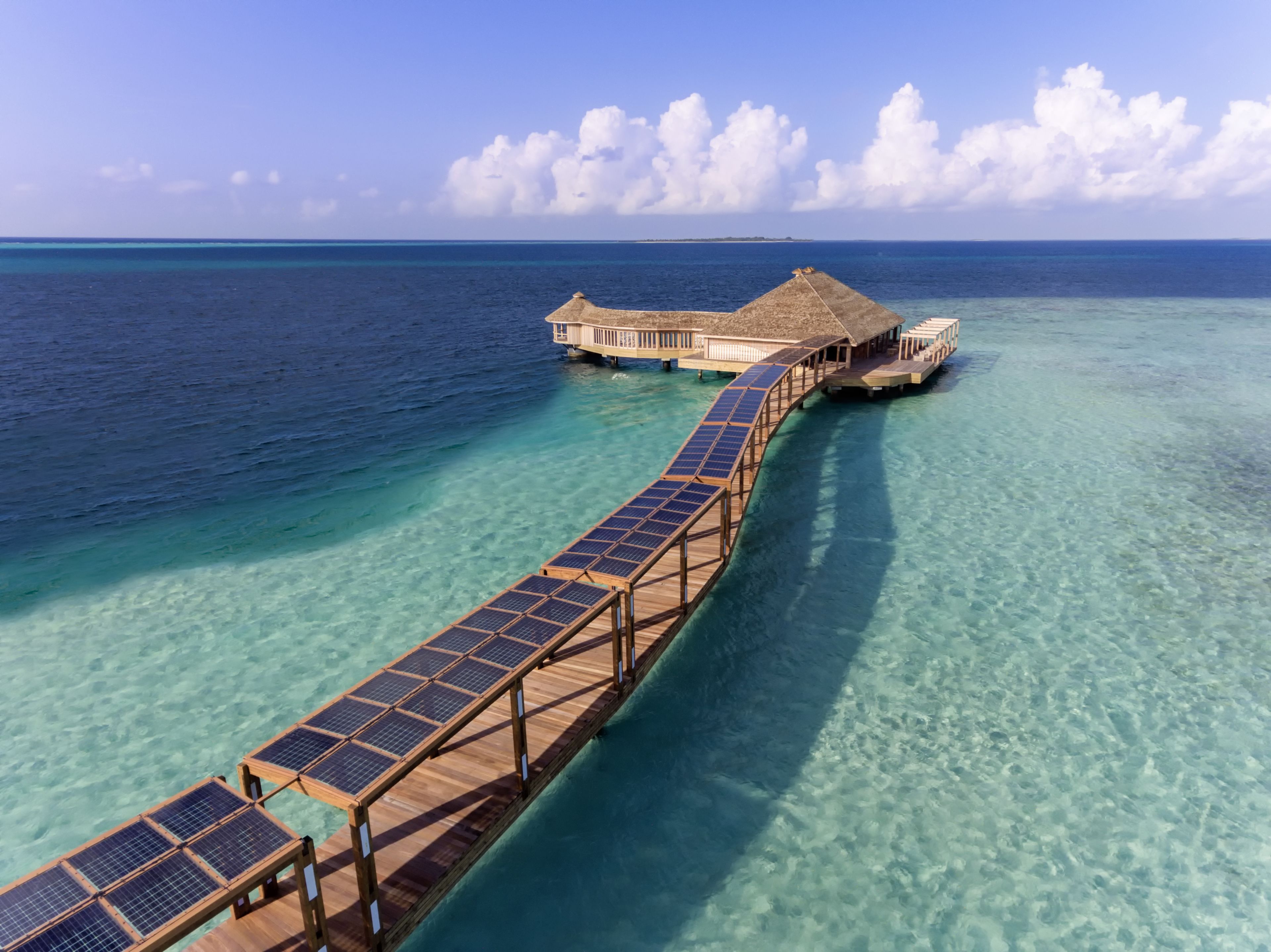 A wooden overwater walkway with solar panels, leading to a thatched-roof building with multiple rooms.