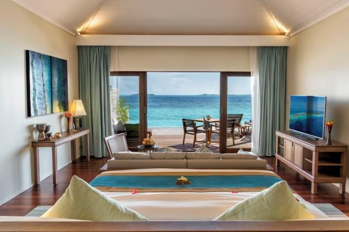 Ein Hotelzimmer mit einem großen Bett, einem Fernseher, einem Essbereich mit Meerblick und einem Balkon mit Tisch und Stühlen.