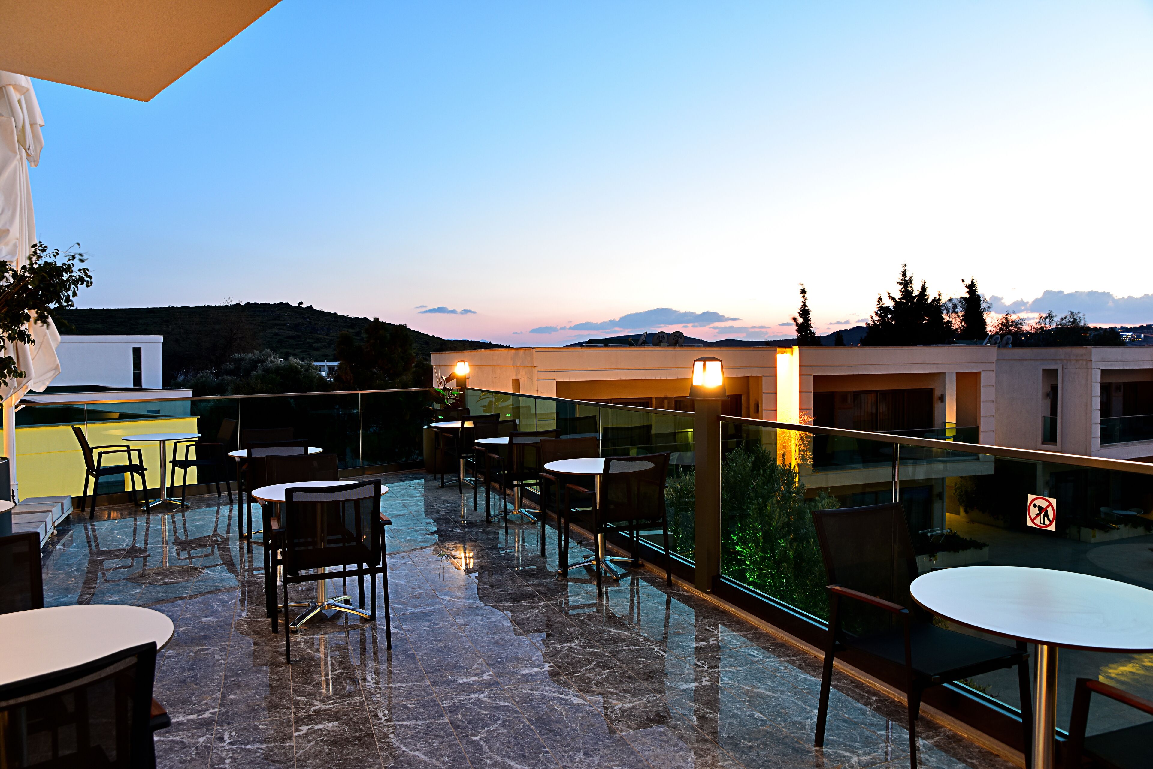 An outdoor terrace with tables and chairs, a glass railing, and a view of the landscape.