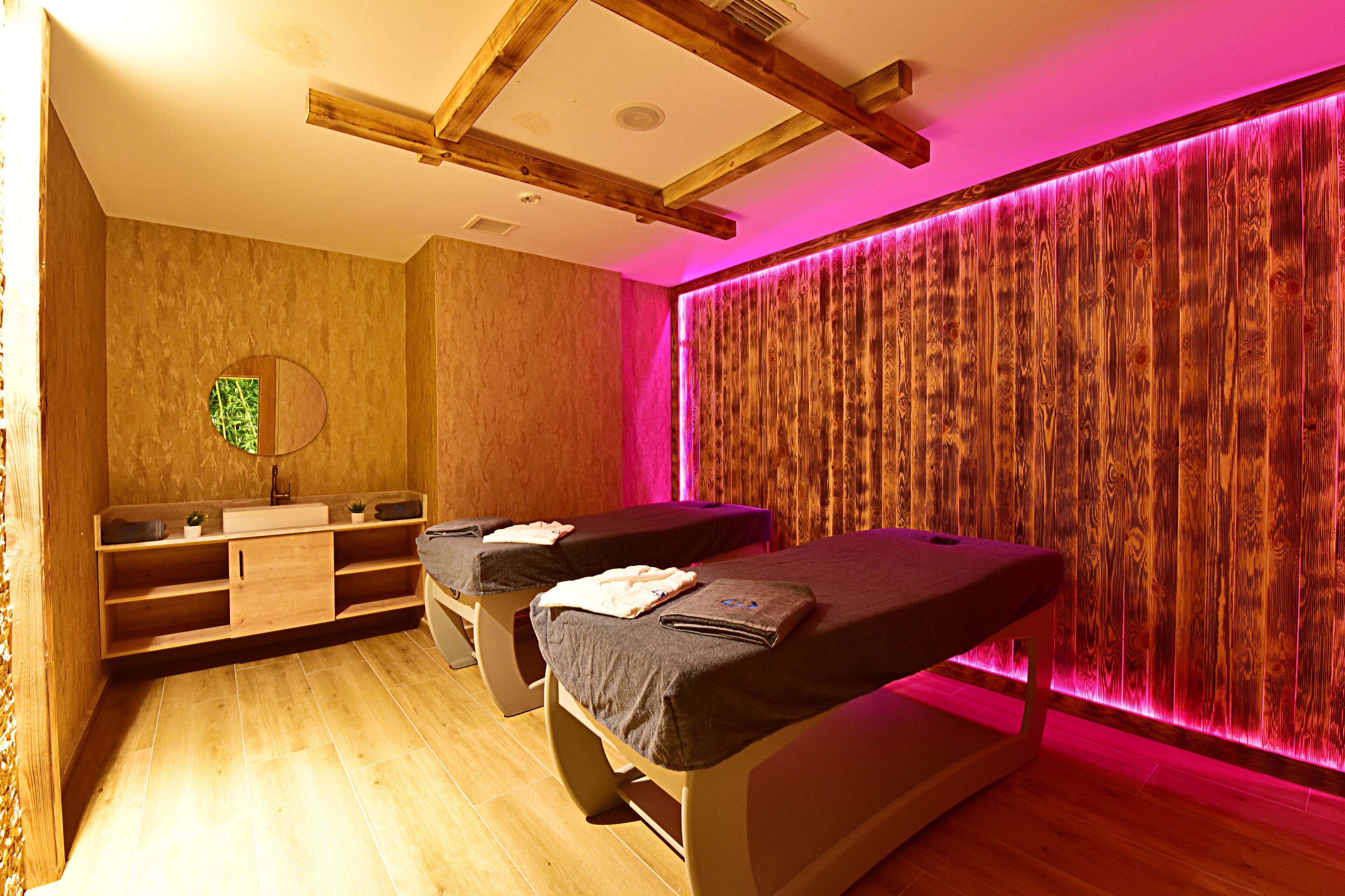 A room with two massage tables, wooden flooring, a wooden wall panel, and a wooden ceiling structure.
