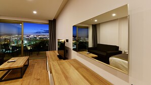 A modern hotel room with a large window offering a city view, a wooden desk, a black sofa, and a bed with a white duvet. - Jasmin Elite Residence & SPA (Bodrum)