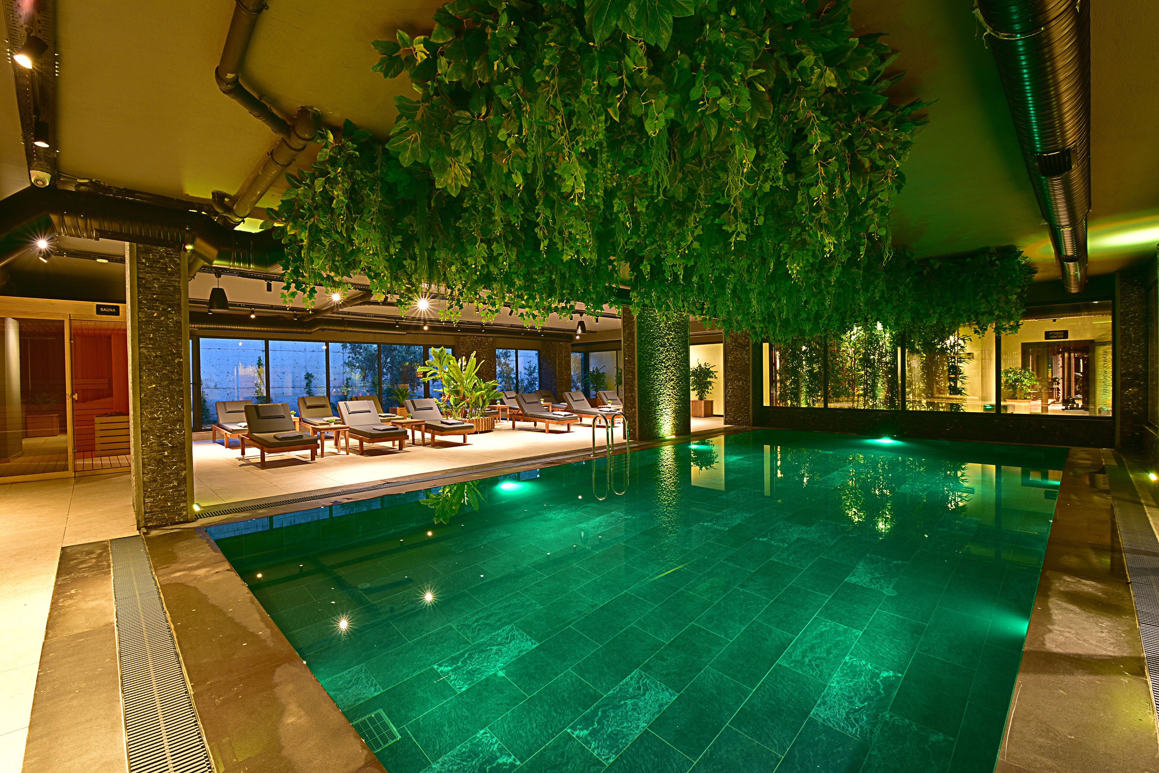 An indoor swimming pool with a large artificial tree, lounge chairs, and a view of the outside at night.