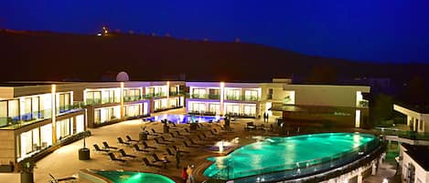 A hotel with a large outdoor swimming pool, multiple lounge areas, and a well-lit building facade.
