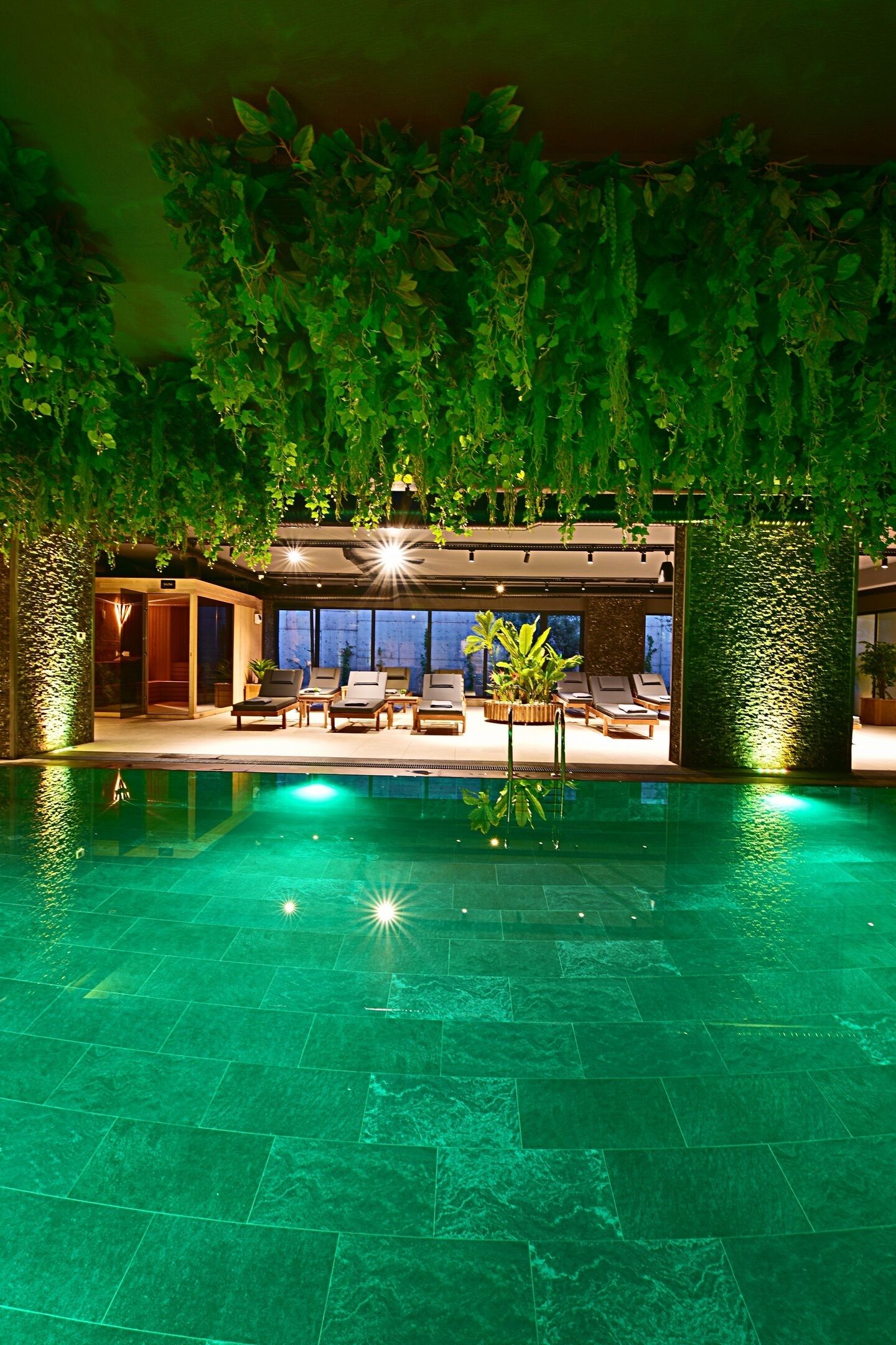An indoor swimming pool with a greenish hue, surrounded by lounge chairs and a wall of hanging plants.