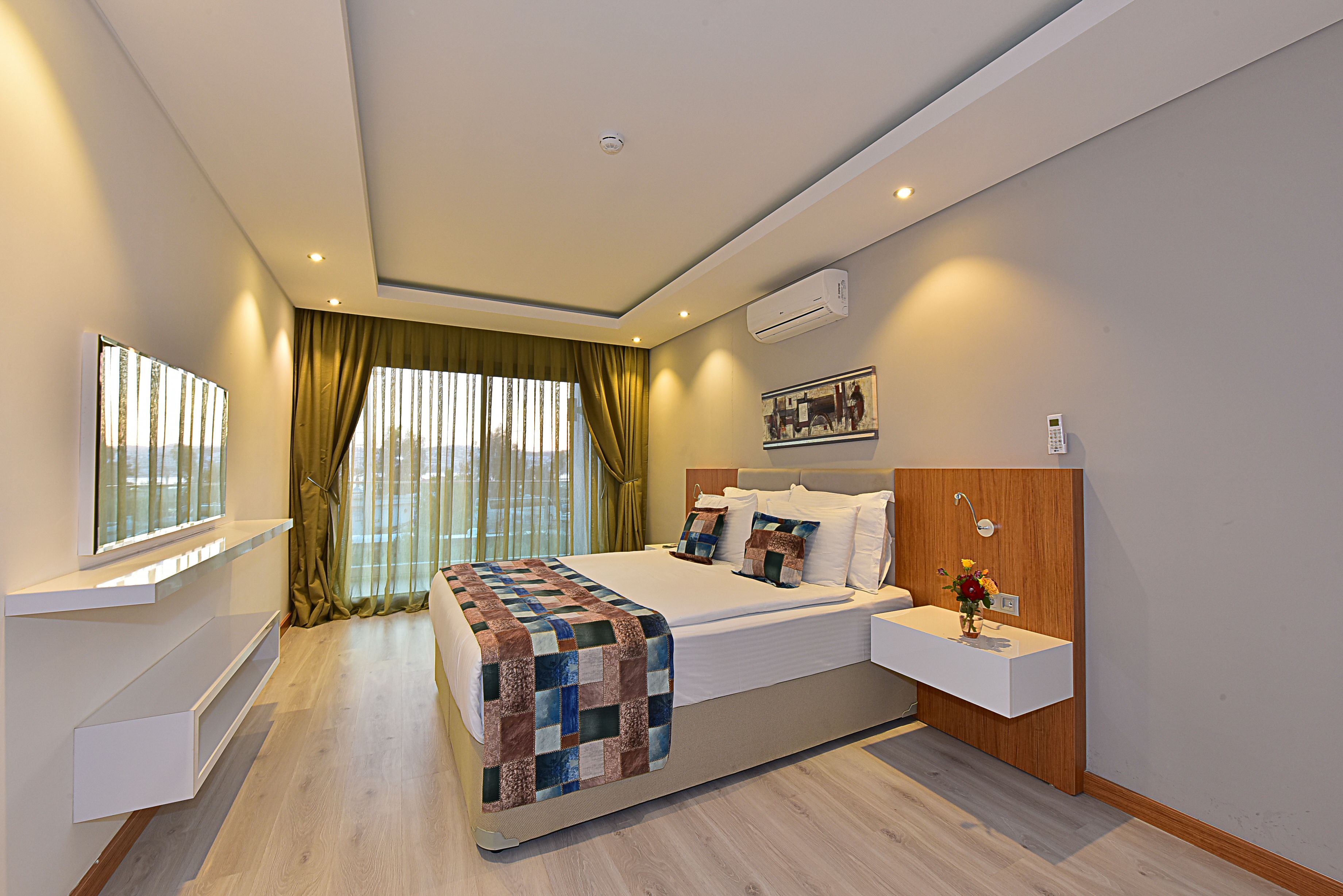 Une chambre d’hôtel moderne avec un grand lit, une table de chevet, une étagère intégrée, une fenêtre avec des rideaux et un climatiseur fixé au mur.