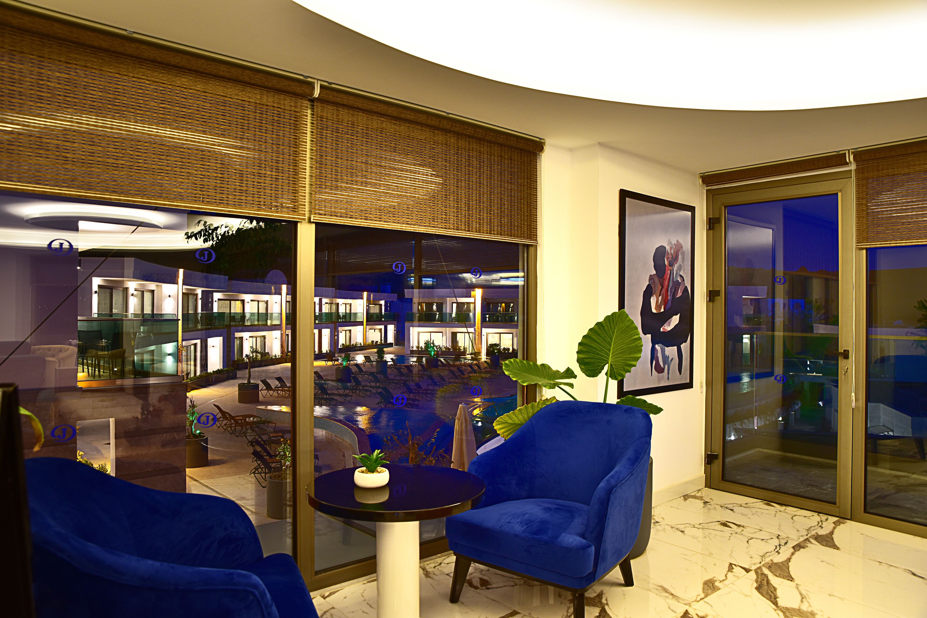 A modern lobby with a curved ceiling, a seating area with blue chairs, a small table with a plant, and a large window with a view of the hotel interior.