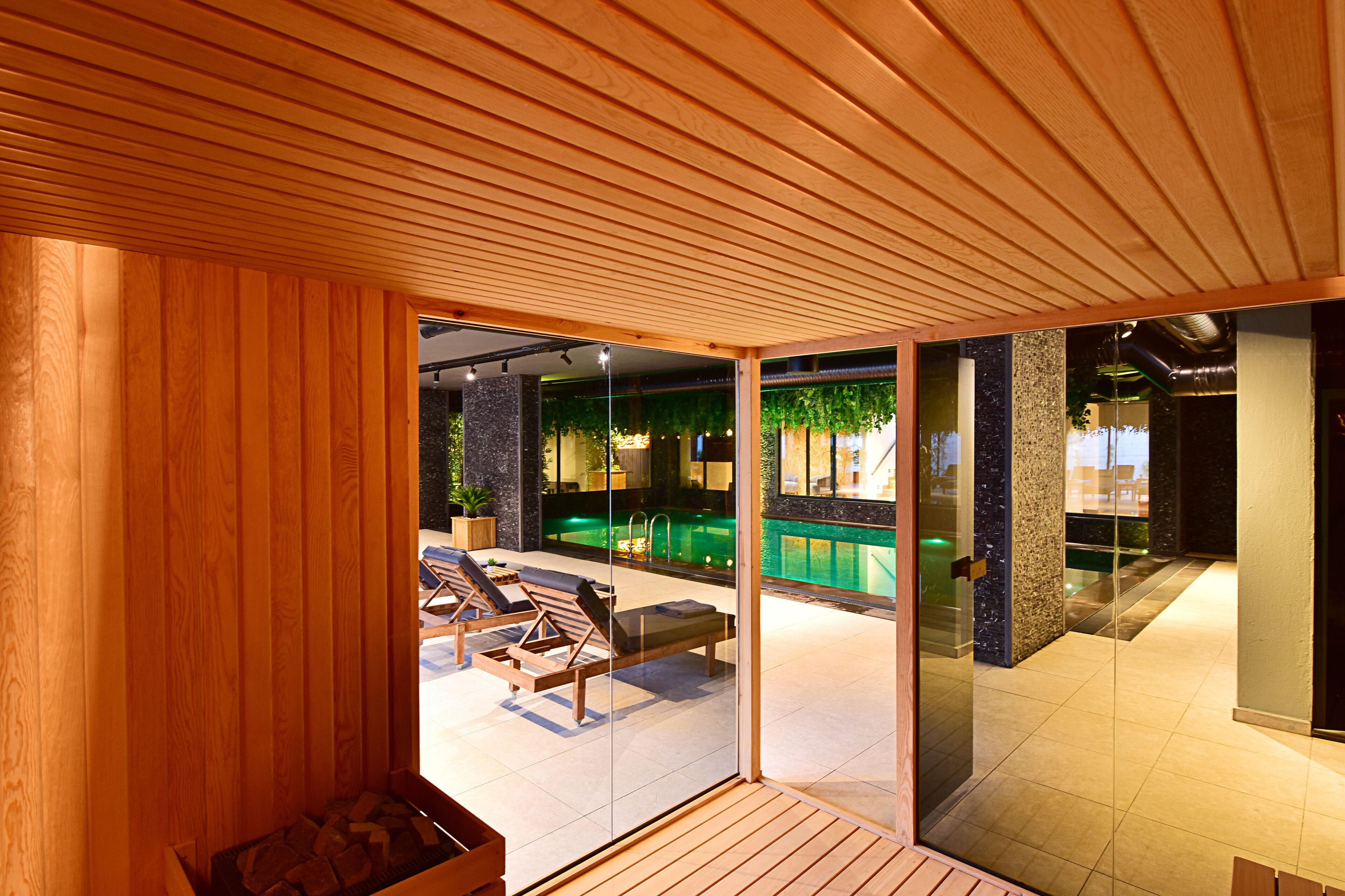 A modern indoor pool area with wooden flooring, lounge chairs, and a glass partition.