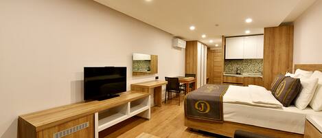 A modern hotel room with a wooden bed, a desk, a television, and a kitchenette.