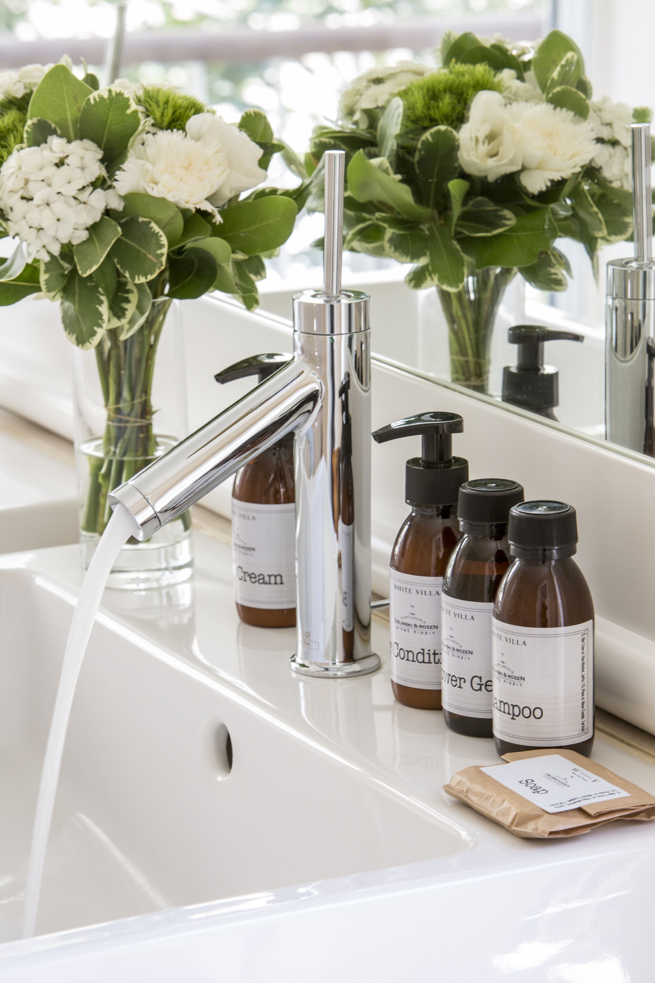 Een wastafel met een chromen kraan, een glazen vaas met witte bloemen en drie flessen haarverzorging van White Villa.