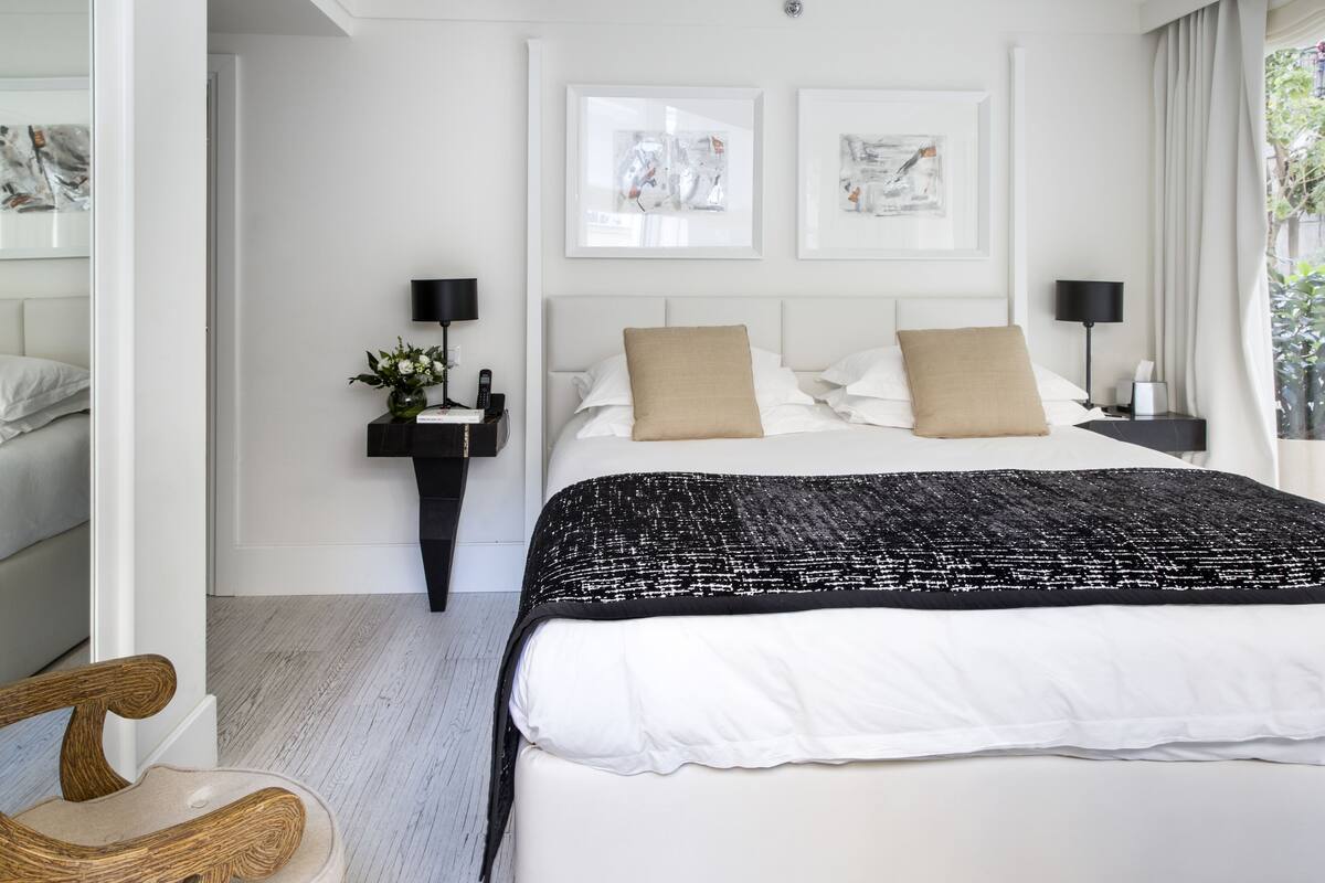 Una habitación de hotel moderna con una cama grande, dos mesitas de noche con lámparas, una mesita auxiliar con un jarrón y obras de arte en las paredes.
