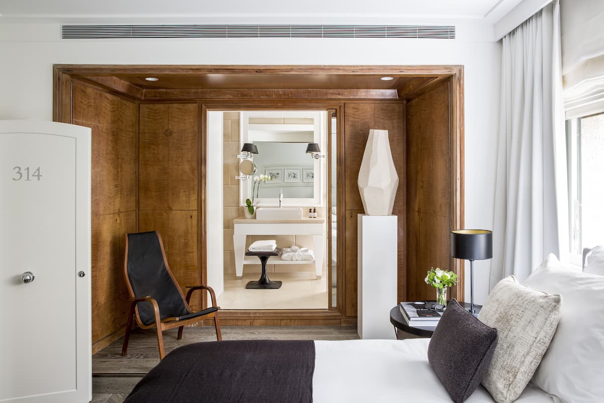 Habitación de hotel con cama, una silla, mesita de noche, lámpara y vista al baño.