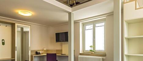 A hotel room with a large bed, a desk, a TV, a window with blinds, and a radiator.