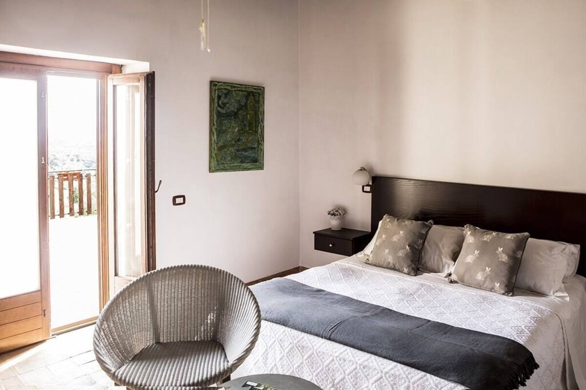 Ein Schlafzimmer mit Bett, Stuhl, Nachttisch, einer Wandlampe und einer Tür, die auf einen Balkon führt.