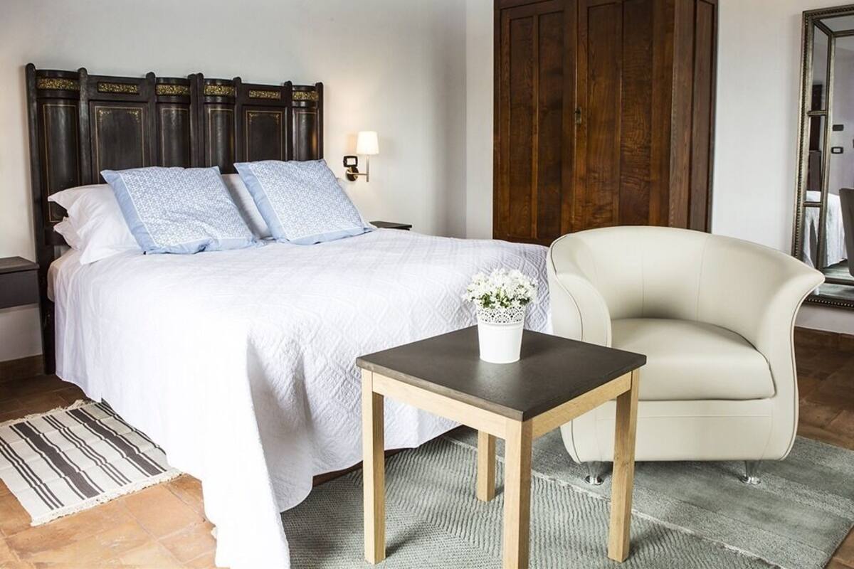Ein Schlafzimmer mit einem Bett, einem Nachttisch, einem Holzschrank, einem Sessel und einem kleinen Tisch mit einer Vase.
