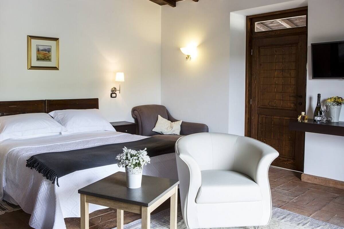 Ein Hotelzimmer mit Bett, einem Sessel, einem kleinen Tisch mit einer Vase, einem an der Wand befestigten Fernseher und einer Holztür.