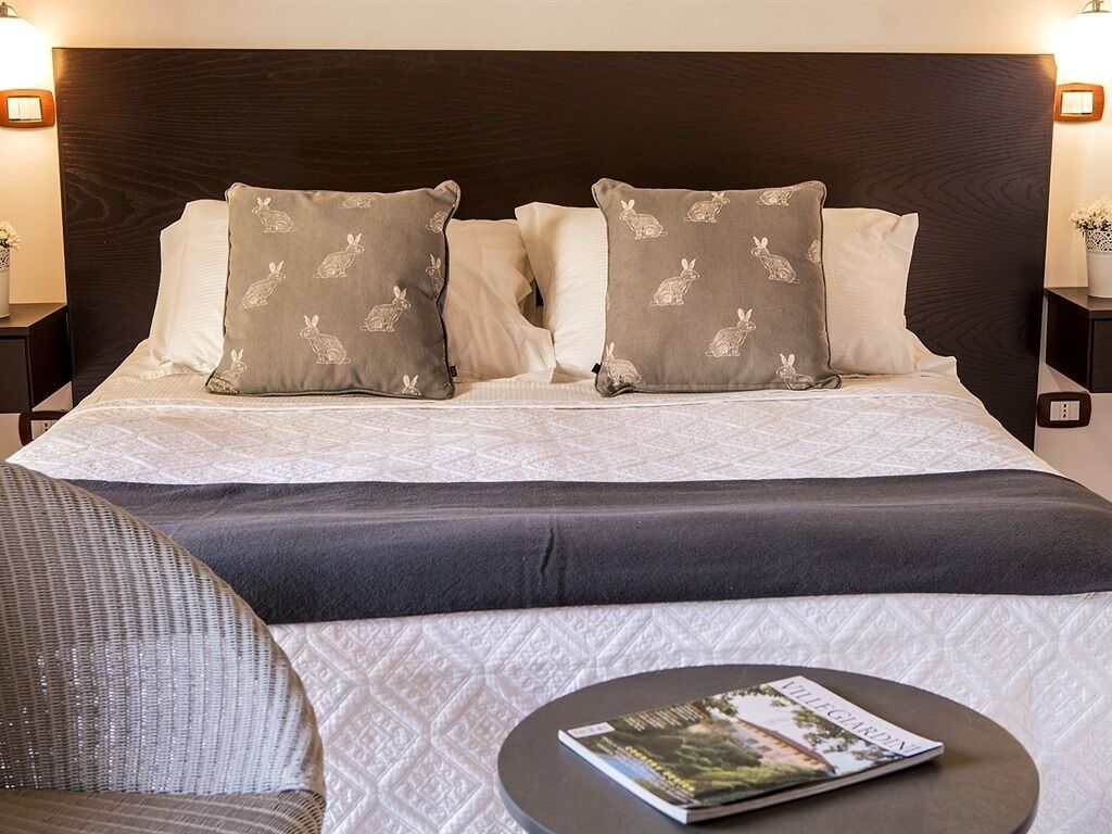 A neatly made bed with patterned pillows, a round bedside table with a magazine, and a dark headboard.