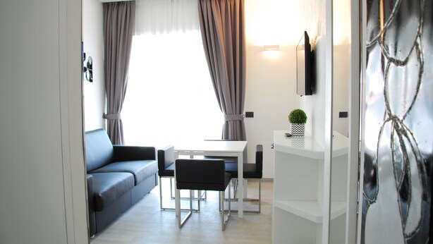 A modern living room with a sofa, a dining table, and chairs. There is a TV mounted on the wall and a small plant on a shelf. - LHP Suite Rapallo (Rapallo)