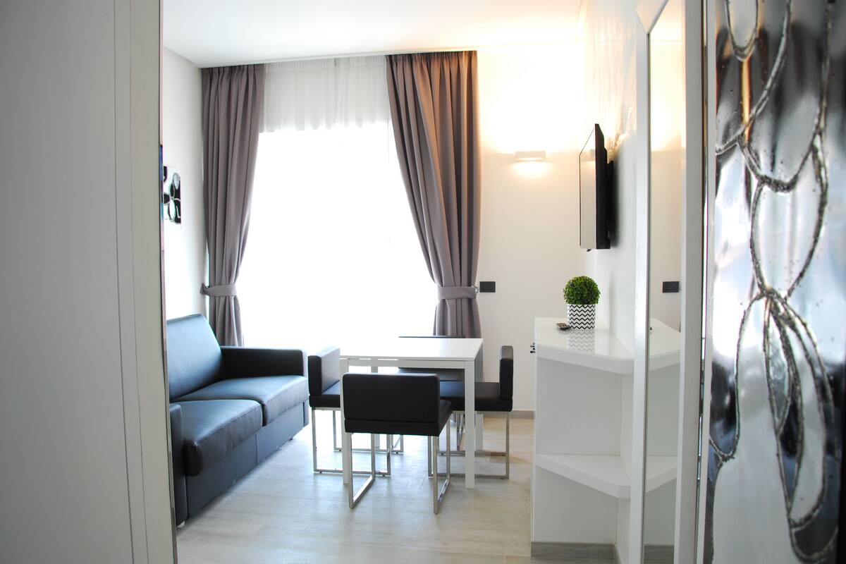 Una sala de estar moderna con un sofá, una mesa de comedor y sillas. Hay un televisor en la pared y una planta pequeña en un estante.