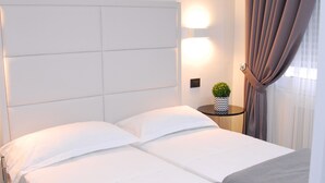 A neatly made bed with white linens and a grey throw blanket, a bedside table with a small potted plant, and a window with curtains. - LHP Suite Rapallo (Rapallo)