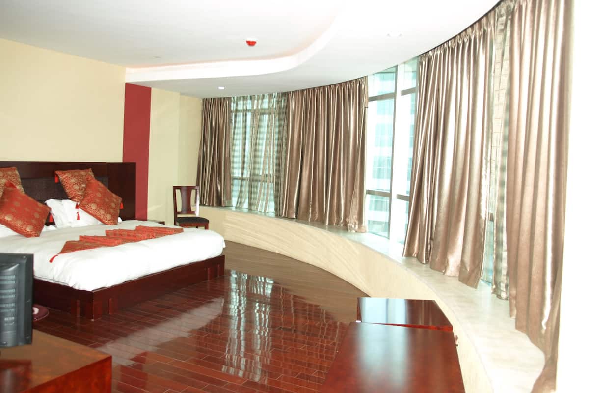 A modern hotel room with a curved wall, a large bed with red and white bedding, a wooden nightstand, and a view of the city through large windows.