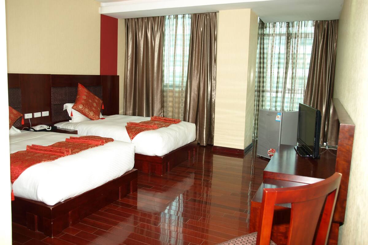 A hotel room with two beds, a desk, a television, and a large window with curtains.