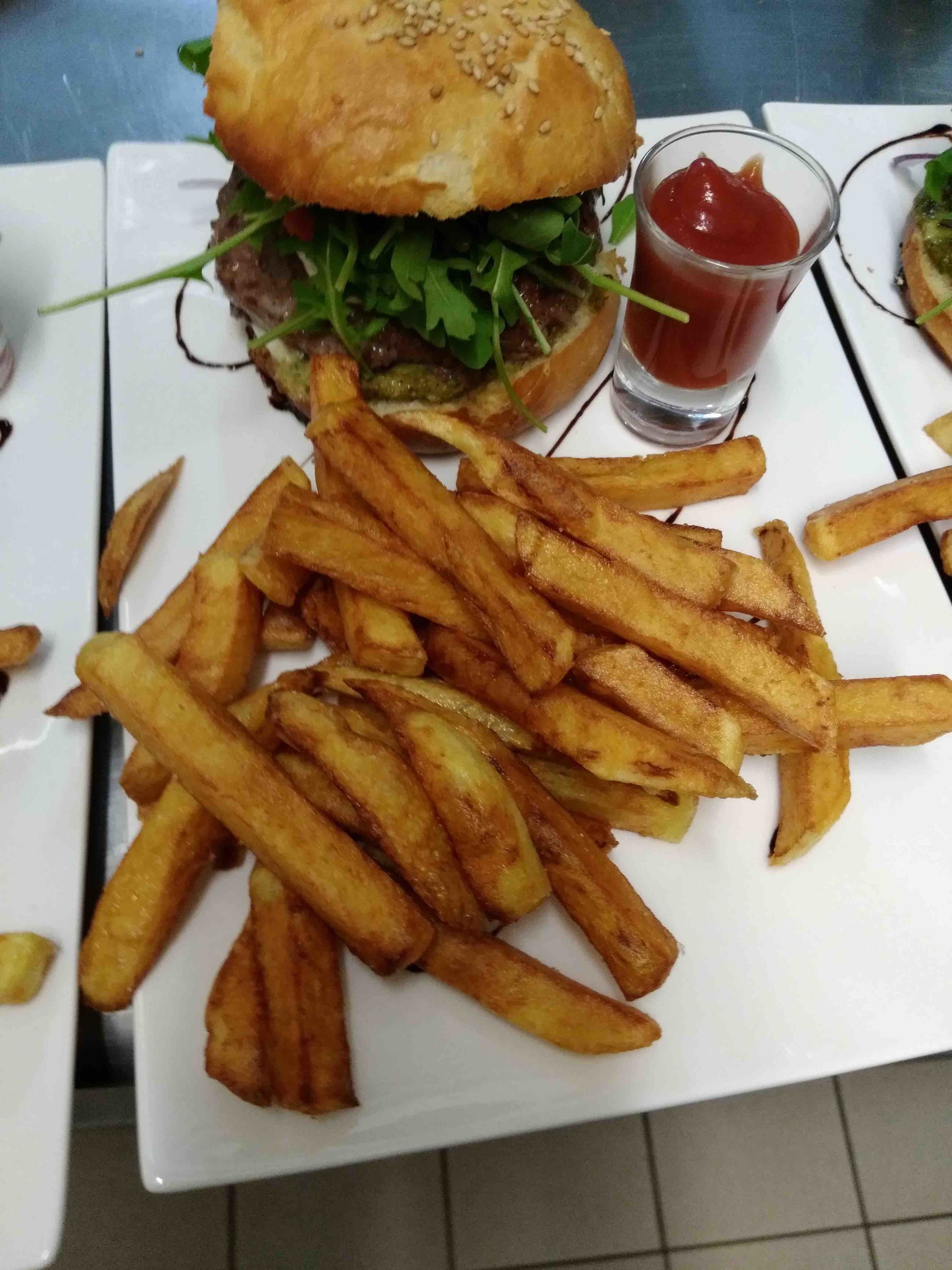 A plate of golden-brown fries and a burger with a sesame seed bun, accompanied by a small glass of ketchup.