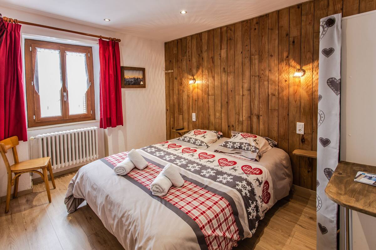 A bedroom with a wooden panel wall, a bed with a red and white checkered bedspread, two wall-mounted lights, a window with red curtains, and a wooden chair.