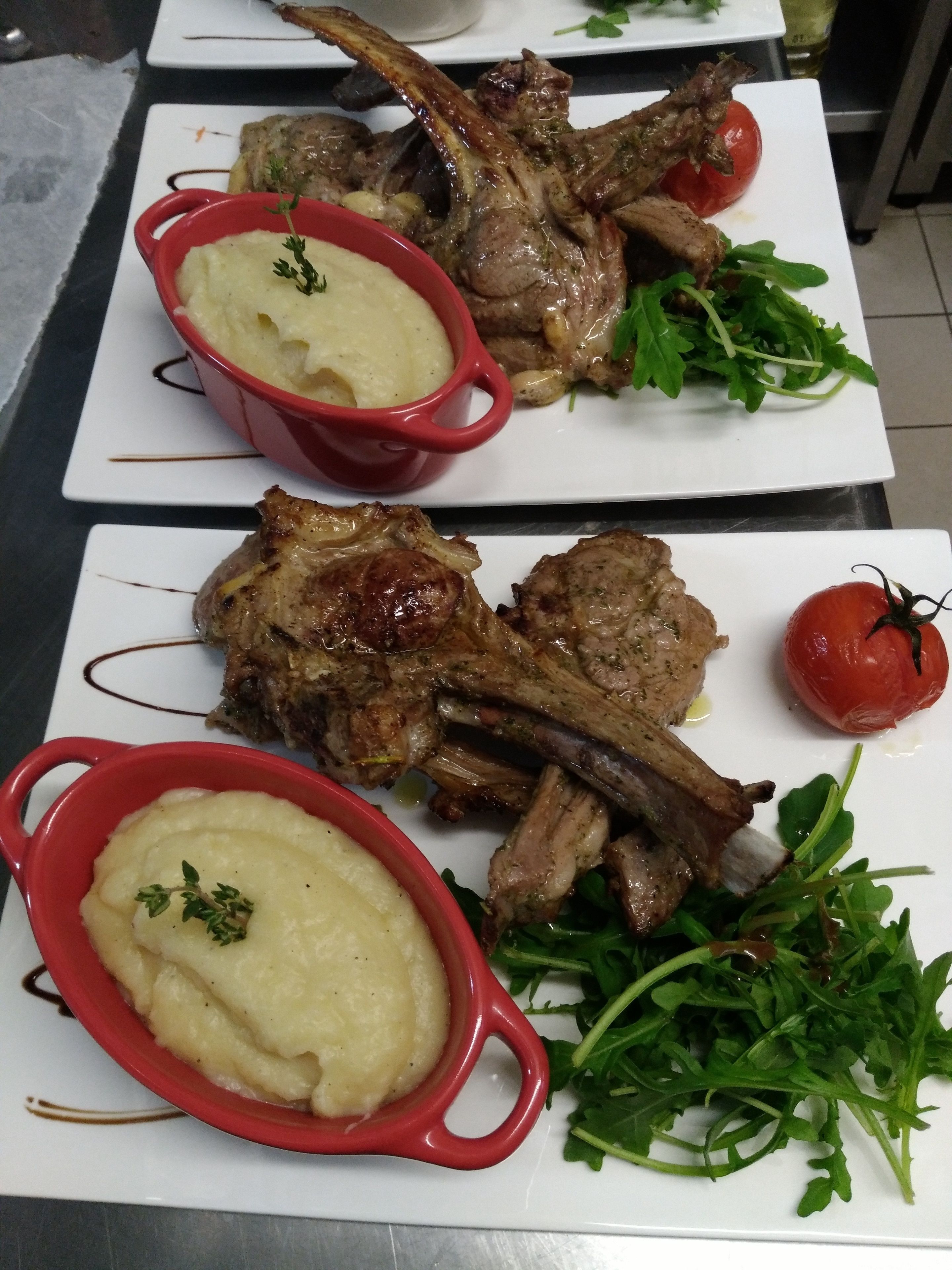 A dining table with two plates of lamb chops, each accompanied by a red bowl of mashed potatoes and a garnish of fresh greens.